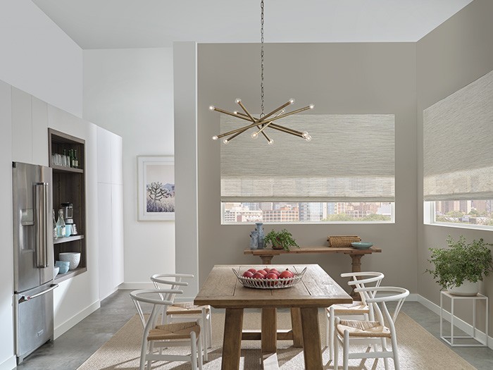 Kitchen area with wooden rectangular table and white wooden chairs with Hunter Douglas Designer Roller Shades and a modern gold overhanging lamp.